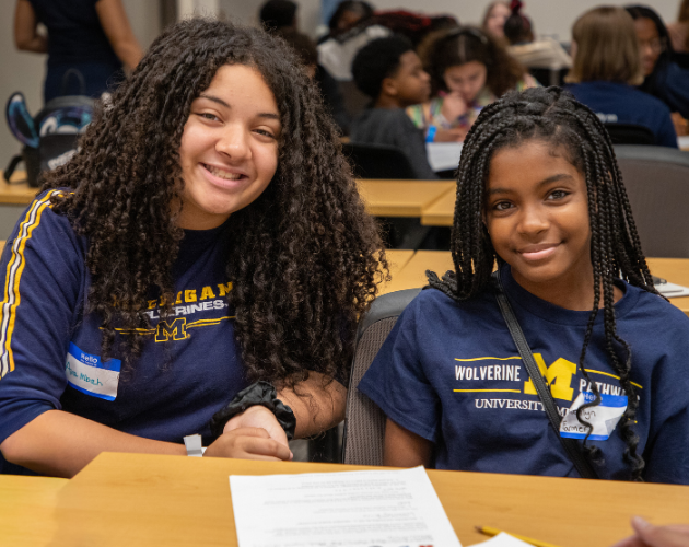 Two students with long braided hair sit at a lecture hall desk, smiling at the camera while working with papers and pens; other students sit in rows behind them.