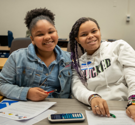 Two people sitting at a classroom table, smiling at the camera while working with printed worksheets, crayons, and a phone; desks and chairs are visible in the background.