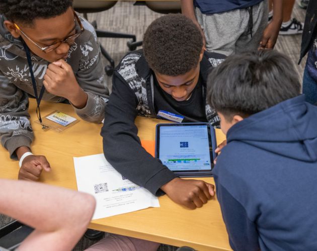 Several students gathered around a wooden table, writing and using laptops with notebooks and papers spread out as they collaborate.
