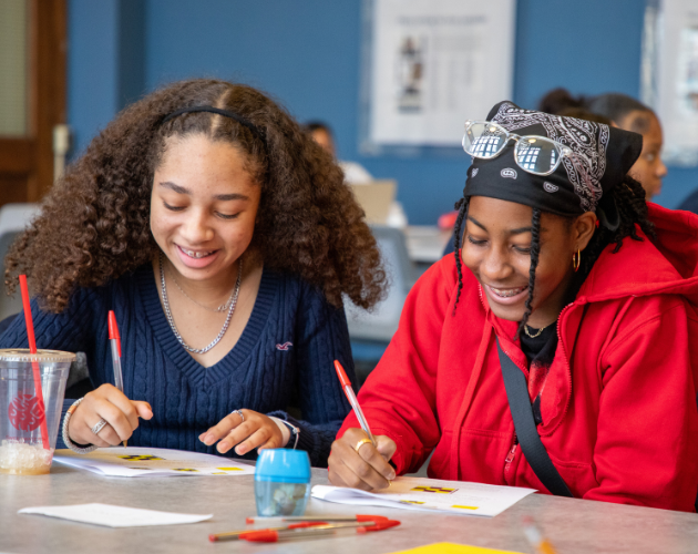 Two students sit at a table drawing and writing with colored markers in a classroom, smiling while working on papers; red and blue drinks sit nearby.