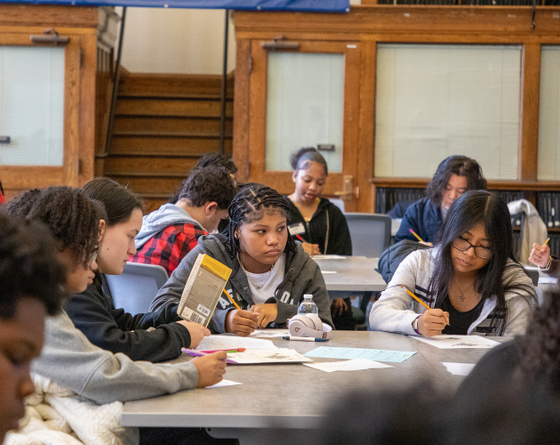 Students sit around a long table working on papers and books in a library classroom, with a blue banner hanging on the wall behind them.