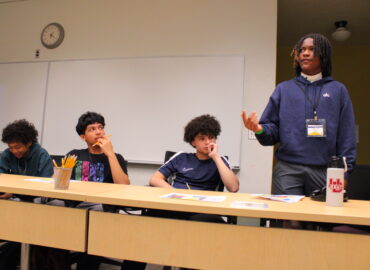 Group of students sitting next to each other in a classroom. One student is standing and speaking to the class.
