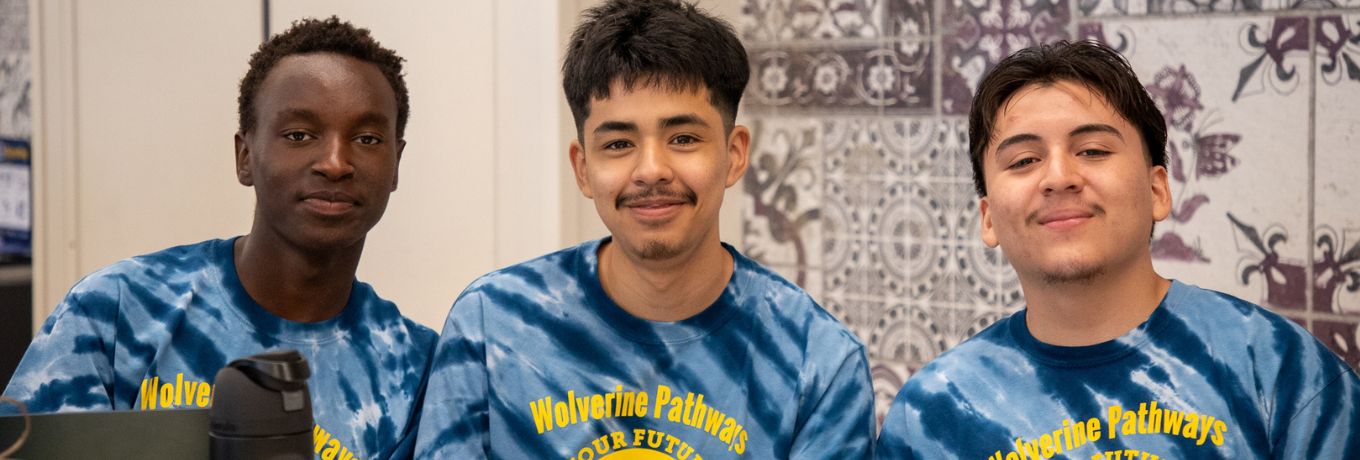 Three students in matching blue “Wolverine Pathways” T-shirts sit behind a table with papers, smiling while a water bottle and pen rest on the tabletop.