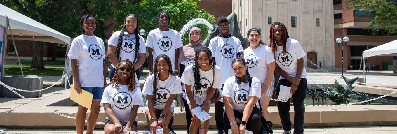 Group photo of students in white T-shirts and shorts, standing and kneeling outdoors on a sunny campus walkway with trees and a tall stone building in the background.