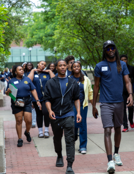 A group of students in casual clothing walk together along a campus walkway lined with trees and brick paths, with benches and stone walls on both sides.