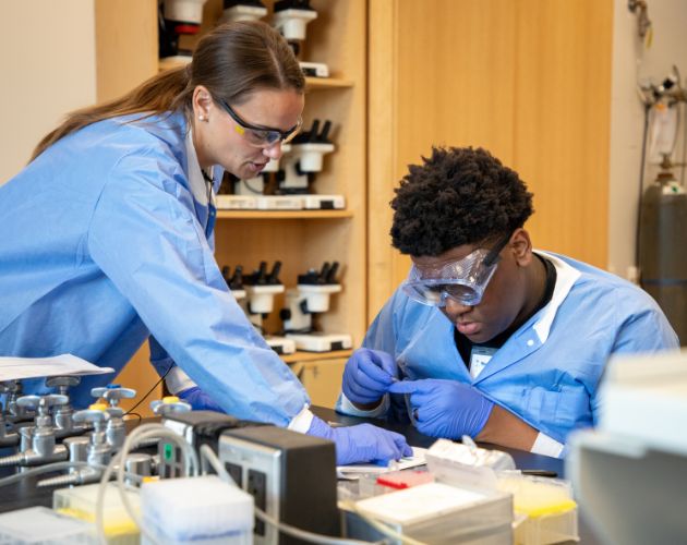 Two people in blue lab coats and safety goggles work at a bench with microscopes and lab equipment, preparing samples in a cleanroom-style laboratory with shelves in the background.