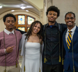 Four young people pose together in a school hallway, smiling at the camera, wearing formal outfits and colorful accents, with a stained-glass window in the background.