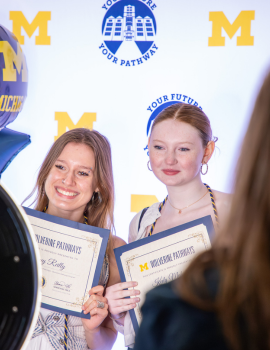 Two people holding certificates and smiling at a university awards backdrop with yellow “M” logos, while another person stands in the foreground and a large camera lens partially blocks the left side.