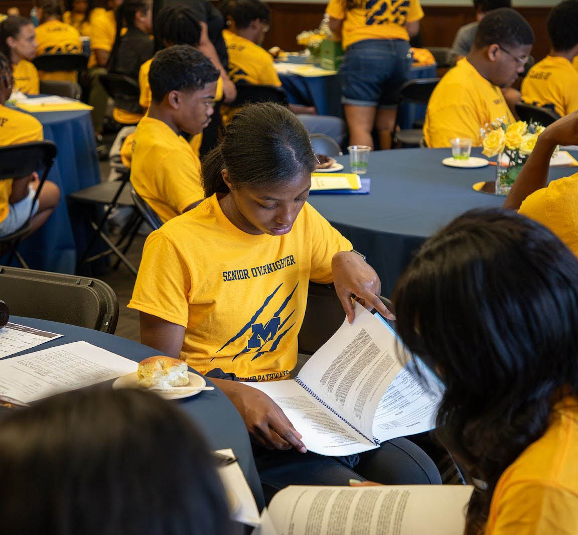 girl sitting at table looking down at book wearing yellow t-shirt