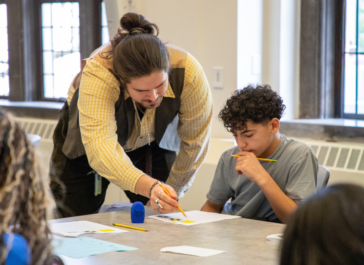 An adult leaning over a table helps a child draw with a yellow pencil, with papers and supplies spread out; a classroom setting with large windows in the background.