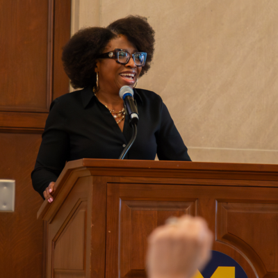 A speaker in a dark blazer stands at a wooden podium with a microphone, addressing an audience in a wood-paneled room; a University of Michigan logo is visible on a nearby attendee’s cap.