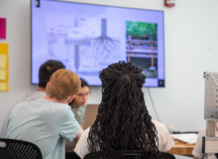 A classroom or office meeting viewed from behind, with people seated at desks watching a wall-mounted screen showing charts and a green image, while a gray filing organizer sits on the right.