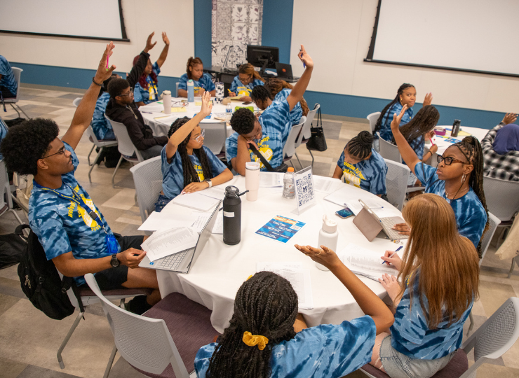 A group seated around a long table in a classroom, eating and talking while raising their hands, with papers, notebooks, and drinks on the table and large projection screens on the walls.