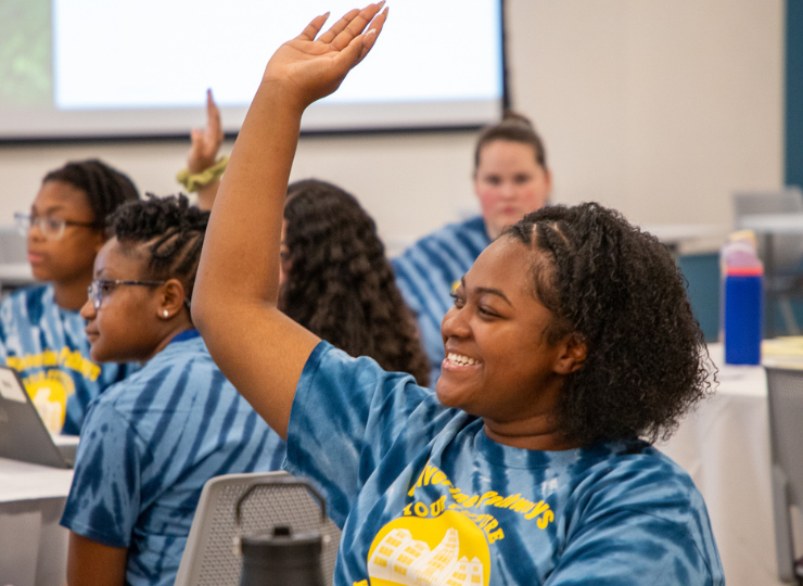 In a classroom, a seated student in a blue shirt raises a hand while classmates listen and a teacher stands near a projection screen.