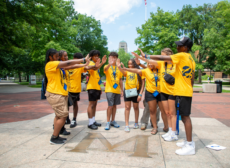 A group of people in matching yellow shirts outdoors in a sunny park, dancing and cheering together on a paved walkway with trees and a tall building in the background.