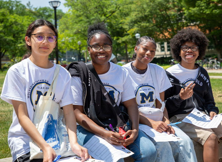 Four students in matching T-shirts sit together on a lawn outdoors, smiling and holding books or papers, with trees and a lamppost in the background.