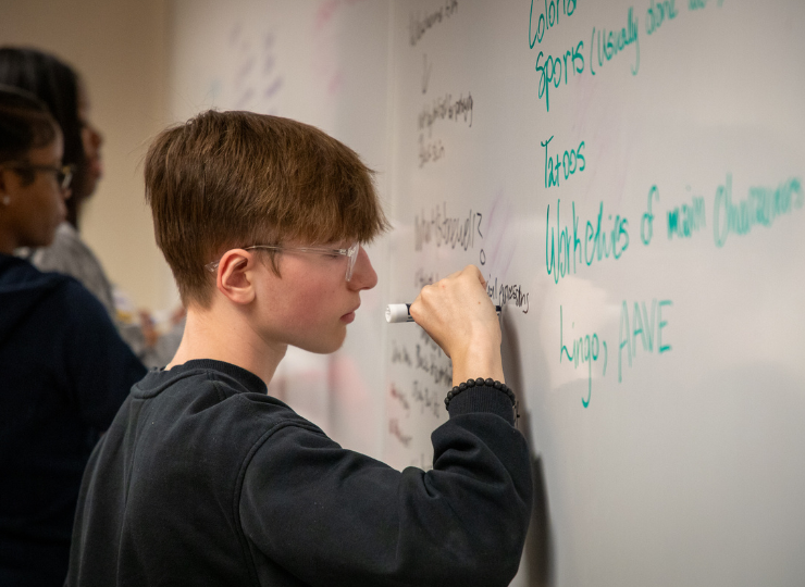 A student in a dark hoodie writes notes on a whiteboard with teal and black text while another student stands behind, suggesting a classroom or study session.