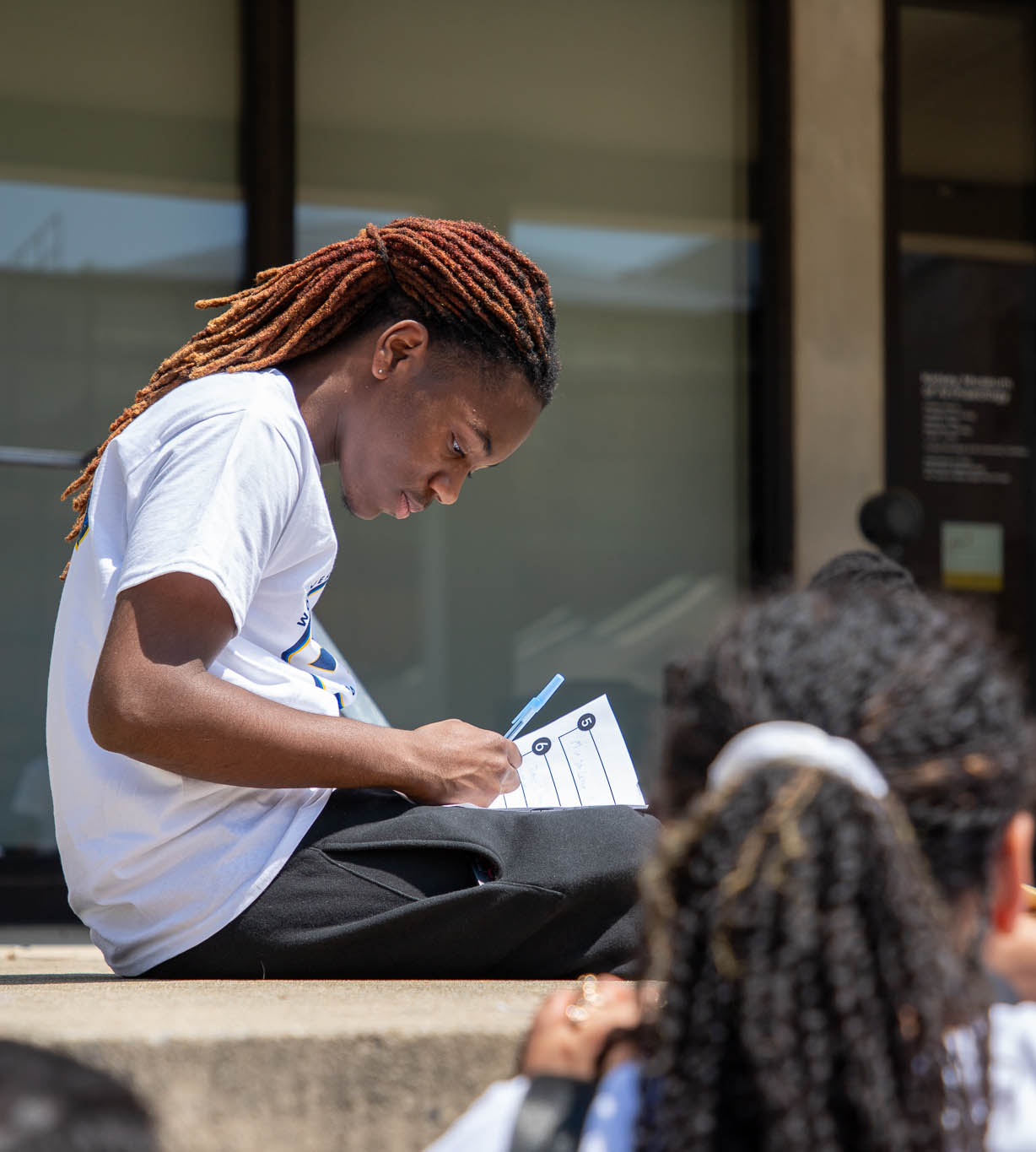 boy with dreadlocks in white shirt sitting outside writing on a paper
