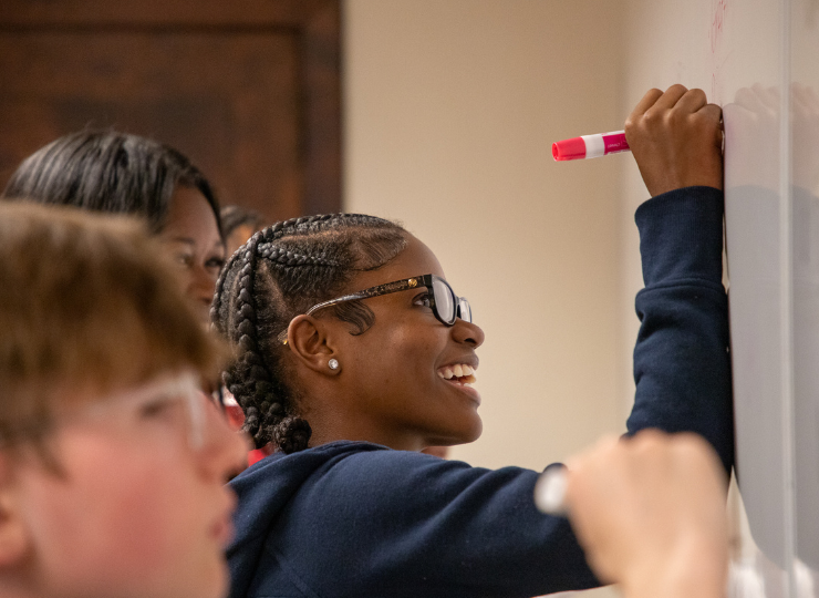 A student with braided hair and glasses writes with a pink marker on a whiteboard while others watch in a classroom setting.