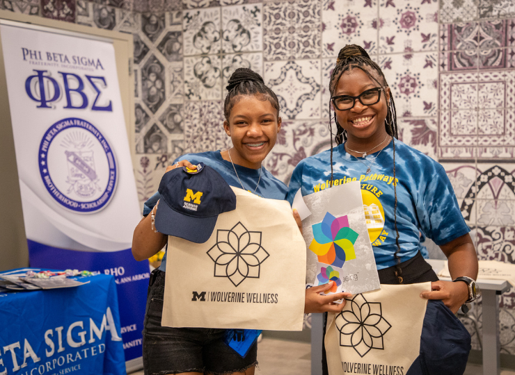 Two students in blue T-shirts stand indoors holding tote bags with printed logos and floral designs, smiling in front of a patterned wall and a Phi Beta Sigma banner.